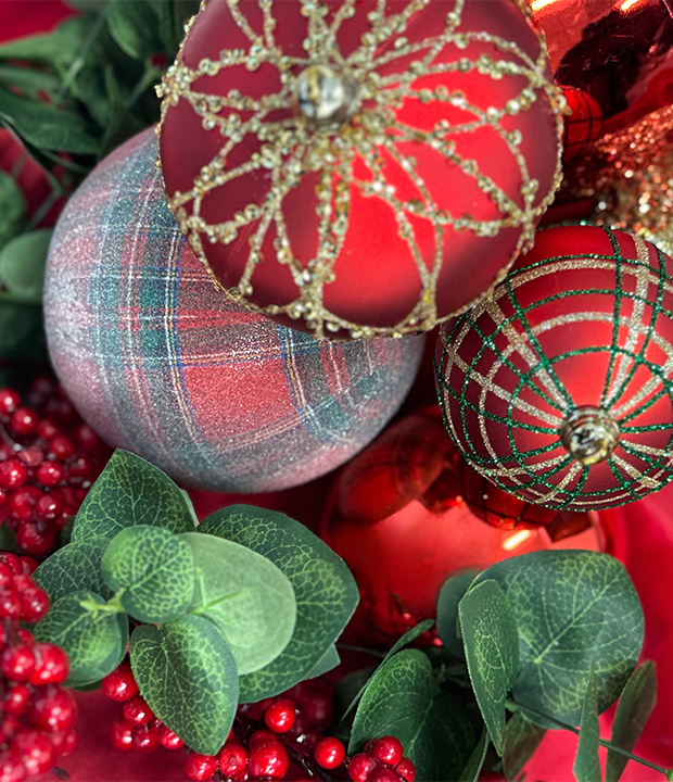 Close-up of Christmas ornaments with red and gold glitter, plaid design, red berries, and eucalyptus leaves.