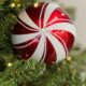 Close-up of a glossy red and white candy cane-patterned Christmas ornament hanging on a green pine branch with soft warm fairy lights.