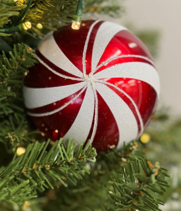Close-up of a glossy red and white candy cane-patterned Christmas ornament hanging on a green pine branch with soft warm fairy lights.