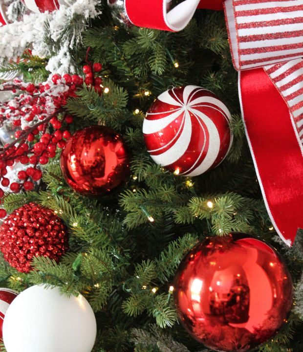 Close-up of a Christmas tree with glossy red baubles, candy-cane striped ornaments, and clusters of red berries among green pine needles and twinkling white fairy lights, creating a festive holiday atmosphere.