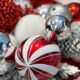 Close-up of Christmas ornaments including a red and white candy cane-patterned bauble, silver and glittery balls, and red ornaments with snowflake patterns, surrounded by pine cones and red glitter.