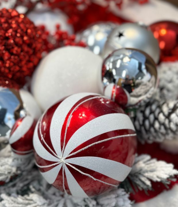 Close-up of Christmas ornaments including a red and white candy cane-patterned bauble, silver and glittery balls, and red ornaments with snowflake patterns, surrounded by pine cones and red glitter.