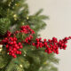 Close-up of a Christmas tree branch with red berries and warm fairy lights among green pine needles, set against an off-white background.