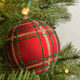 Close-up of a red plaid Christmas ornament with green and gold glitter stripes hanging on a tree, with pine needles and soft holiday lights in the background