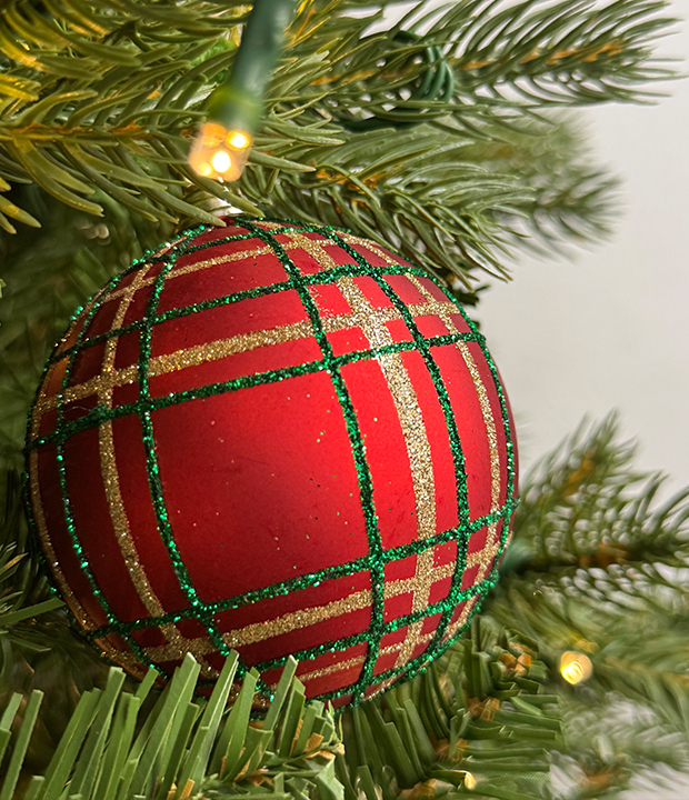 Close-up of a red plaid Christmas ornament with green and gold glitter stripes hanging on a tree, with pine needles and soft holiday lights in the background