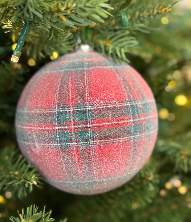 Close-up of a round red and green plaid Christmas ornament hanging on a tree, with soft texture, warm festive colors, and pine needles in the background.