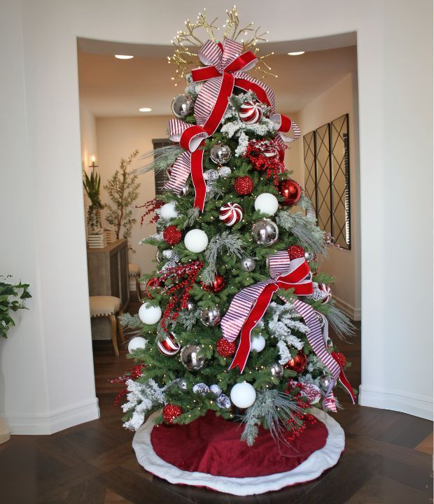 Christmas tree with red and white ribbons, silver and red ornaments, and a red bow, in a modern living room with wooden floors and white walls.