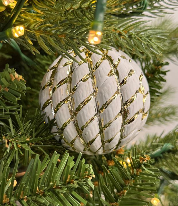 Close-up of a white Christmas ornament with gold zigzag patterns nestled in green pine needles, with warm twinkling lights creating a festive holiday atmosphere.