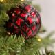 Close-up of a glossy red Christmas ornament with black diamond-patterned texture, nestled among green pine needles and warm white fairy lights, creating a festive holiday atmosphere.