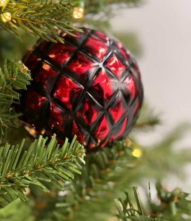 Close-up of a glossy red Christmas ornament with black diamond-patterned texture, nestled among green pine needles and warm white fairy lights, creating a festive holiday atmosphere.