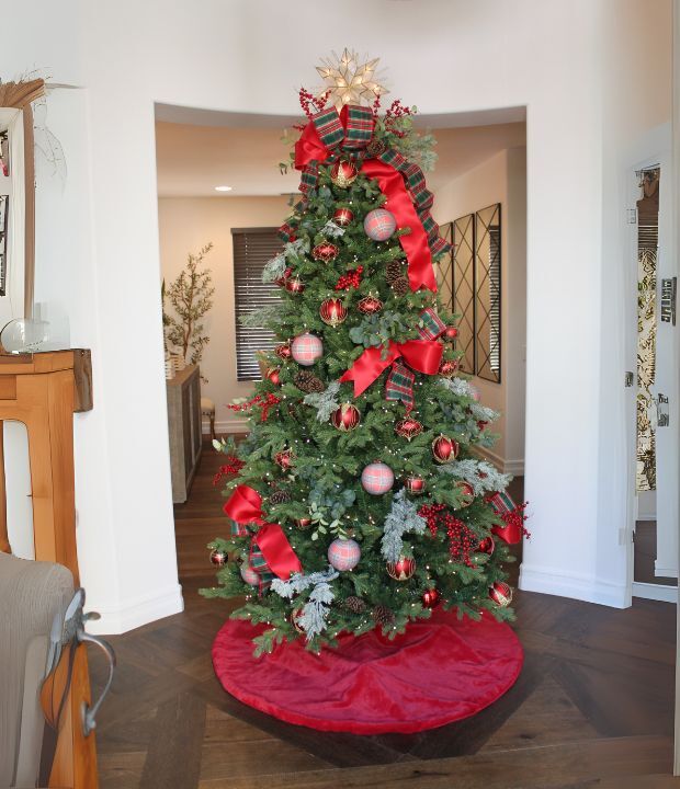 Decorated Christmas tree with red and silver ornaments, ribbons, and bow in a modern living room with wooden floor, white walls, and warm lighting.