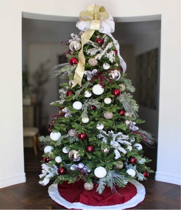 Large green Christmas tree decorated with white, silver, and red ornaments, pine cones, faux snow, and a white and gold bow, standing on a burgundy velvet skirt in a bright, festive holiday setting.